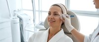 Young Caucasian woman receiving professional facial treatment at modern beauty clinic with dermatologist applying skincare procedure for anti-aging wellness.