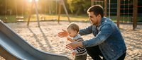 Young Caucasian father in denim jacket helping his toddler son slide down playground equipment during golden hour sunset at outdoor park.