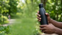 Man with dark skin tone holding black reusable water bottle outdoors in green park setting, promoting eco-friendly hydration and sustainable lifestyle choices.