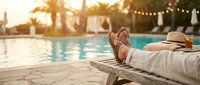 Relaxing poolside vacation scene with legs on lounge chair, tropical palm trees and string lights creating warm sunset ambiance for travel and leisure content.