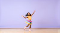 Young girl with dark hair and medium skin tone wearing a bright yellow top and colorful rainbow tutu, dancing joyfully with arms raised on wooden floor against purple background for children's activity and movement content.