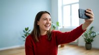 Young Caucasian woman in red sweater taking selfie with smartphone, smiling happily in bright modern home interior with plants and natural light.