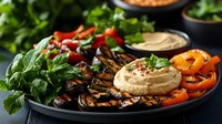 Middle Eastern appetizer platter with creamy hummus, grilled eggplant, fresh herbs, roasted bell peppers, and garnishes served on dark plate against moody background.