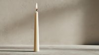 Elegant cream colored candle with bright flame burning against neutral beige wall background creating warm ambient lighting for home decor and relaxation.