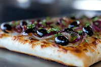 Fresh baked flatbread topped with black olives, red onions and herbs, macro view showing crispy crust and melted cheese with selective focus.