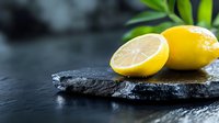 Fresh yellow lemon cut in half on dark slate stone surface with green leaves and water drops for culinary cooking and healthy nutrition concepts.