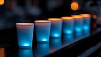 Glowing blue cocktails in white paper cups create atmospheric bar lighting effect with orange bokeh background, modern party drink presentation.