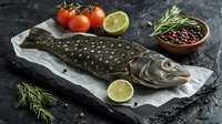 Fresh raw pike fish on paper with lime, cherry tomatoes, rosemary sprigs and peppercorns in wooden bowl on black stone background. Food preparation concept.