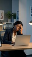 Stressed African American businesswoman with headache working late at laptop in modern office environment for workplace wellness concepts.