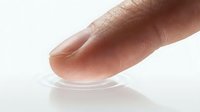Close-up of human finger touching clear contact lens on white surface showing optical vision correction technology for eyecare and medical use.