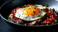 Savory rice bowl topped with fried egg, glazed meat, red sauce, sesame seeds, and green onions, served in a dark ceramic bowl.