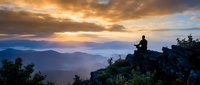 Person meditating on mountain cliff during golden hour sunrise with layered hills and dramatic cloudy sky creating peaceful spiritual moment for wellness content.