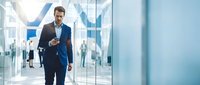 Confident businessman in navy suit walking through modern glass office corridor while using smartphone for corporate communication and business networking.