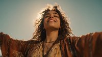 Young mixed-race woman with curly hair spreading arms wide in joyful celebration against golden sunset sky, expressing freedom and happiness outdoors.