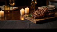 Grilled beef steaks with herbs on wooden board in elegant candlelit dining setting with wine glasses for romantic dinner or fine dining restaurant menu.