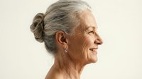 Senior woman with gray hair in bun smiling in profile showing natural aging and confidence against neutral background for lifestyle content.