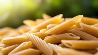Uncooked penne pasta scattered on wooden surface with soft natural lighting and shallow depth of field creating appetizing food composition.