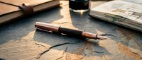 Fountain pen with wooden accents rests on textured slate surface surrounded by open books and ink bottle, evoking writing, literature, and creative workspace ambiance.