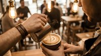 Professional barista creating latte art in coffee cup with steamed milk, warm cafe atmosphere with vintage Edison bulbs and blurred customers in background.