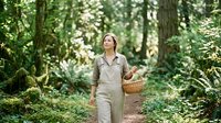 Young woman with light skin tone and brown hair, wearing beige overalls and a collared shirt, carrying a wicker basket while walking through a lush forest path surrounded by green trees and dappled sunlight.