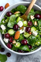 Fresh spinach salad with broccoli, cucumber, cranberries, feta cheese and almonds in white ceramic bowl. Healthy vegetarian lunch with wooden spoon.