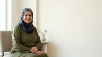 Middle-aged woman wearing olive green hijab sitting in modern interior space with neutral background, suitable for diversity, healthcare, lifestyle and portrait projects.