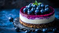 Fresh blueberry cheesecake with vibrant purple layer and cookie crust garnished with ripe berries and mint leaves on dark background, food photography.