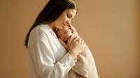 Young Caucasian mother tenderly holds sleeping newborn wrapped in soft blanket against warm beige background, capturing love and maternity.