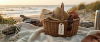 Wicker picnic basket with summer tag, straw hat, and blanket on sandy beach at golden sunset, ocean waves in background.