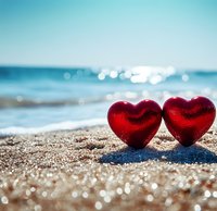 Two red heart decorations on sandy beach with sparkling ocean and blue sky background, symbolizing romance and love by the sea.