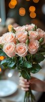 Delicate peach roses bouquet with fresh buds and green leaves held against soft bokeh lights background, creating romantic wedding or celebration atmosphere.