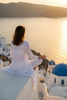 Woman in white clothing meditating at sunset overlooking sea and traditional white buildings with blue domes for wellness and mindfulness concepts.