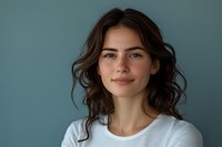 Young Caucasian woman with natural wavy dark hair and minimal makeup wearing white t-shirt smiling confidently against blue background, expressing genuine happiness.