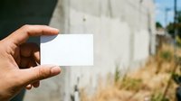 Hand holding blank white business card with blurred concrete wall and natural outdoor background, suitable for branding mockups and corporate identity presentations.