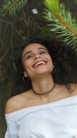 Young woman with curly dark hair laughing joyfully under pine branches, wearing white off-shoulder top and choker necklace, radiating happiness and natural beauty.