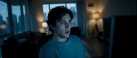 Young man with dark hair and light skin, wearing a blue t-shirt, sitting indoors at night with city skyline visible through windows behind him, looking concerned or worried.