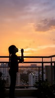 Silhouetted female child blowing soap bubbles on urban balcony at golden sunset, cityscape backdrop with warm sky tones, leisure and childhood moments.