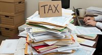Overwhelmed office worker managing massive pile of tax documents and paperwork during busy accounting season with cardboard storage boxes.