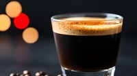 Fresh espresso shot with golden crema in glass cup against dark background with blurred bokeh lights, close up view of aromatic coffee drink.