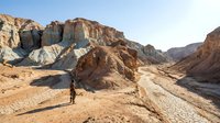 Young male hiker in casual attire exploring vast desert landscape with dramatic rocky formations and canyon terrain under clear blue sky, suitable for travel and adventure content.