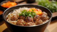 Asian meatball noodle soup with fresh herbs and vegetables in dark bowl. Traditional comfort food with sesame seeds and cilantro garnish for restaurant menu.