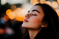 Young Asian woman with closed eyes and serene expression against blurred golden bokeh lights at sunset, side profile portrait capturing peaceful moment of mindfulness.