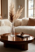 Cozy living room interior with dried pampas grass in crystal vase on round wooden coffee table, white sheepskin sofa with fluffy pillows, natural light.
