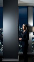 Professional businesswoman in black suit standing by floor-to-ceiling windows overlooking illuminated city skyline at night in modern corporate office.