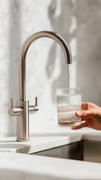 Modern brushed nickel kitchen faucet with water flowing into clear glass held by female hand with manicured nails against neutral marble background.
