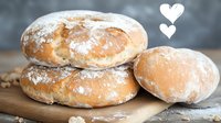 Fresh homemade bread rolls dusted with flour stacked on wooden cutting board against dark background, close up view of crusty artisan baked goods.