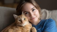 Young Caucasian woman in blue sweater cuddling with ginger cat on grey couch, warm natural lighting creates cozy atmosphere for pet lifestyle and companionship concept.