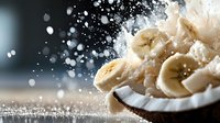 Fresh banana slices on white coconut with splashing milk drops and powder creating dynamic food composition against dark background. Healthy breakfast concept.