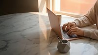 Professional woman working on laptop at marble table with warm sunlight streaming through window creating productive home office atmosphere.
