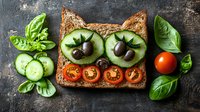 Creative cat face sandwich made with whole grain bread, cucumber slices as eyes, olives as pupils, cherry tomatoes forming smile, garnished with fresh basil leaves.